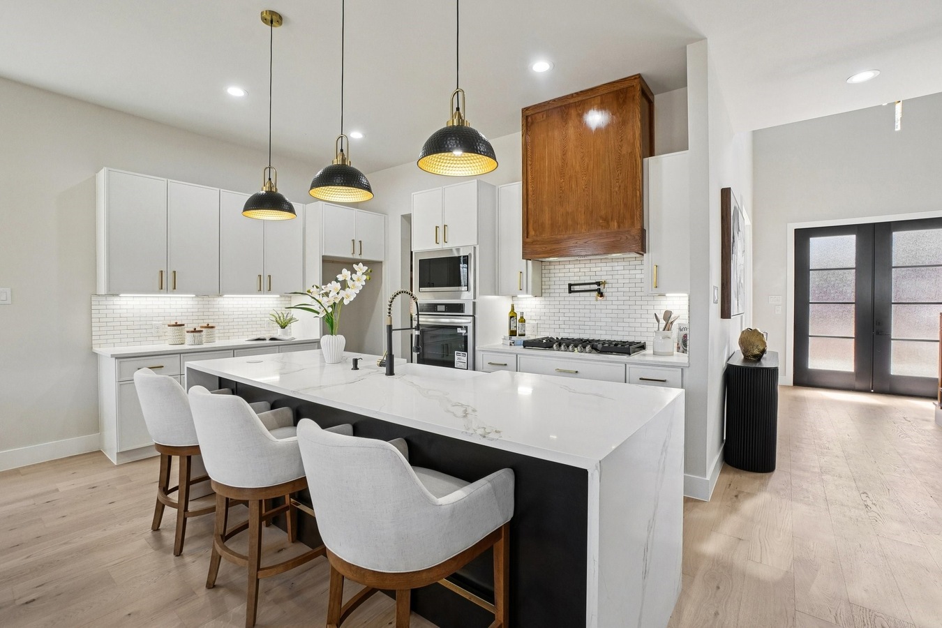 Modern kitchen featuring a large white center island, crisp white shaker base cabinets, contrasting warm wood upper cabinets and range hood cover, black pendant lighting, and stainless steel appliances.