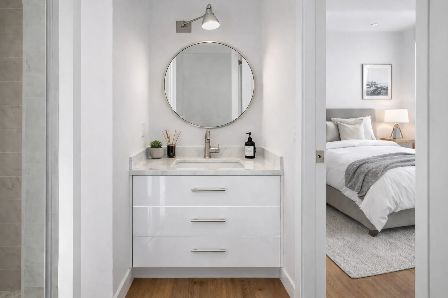 Bright modern bathroom featuring a custom glossy white single vanity with storage drawers, silver hardware, a white countertop, a round mirror, and a wall sconce, situated in a nook adjoining a bedroom.