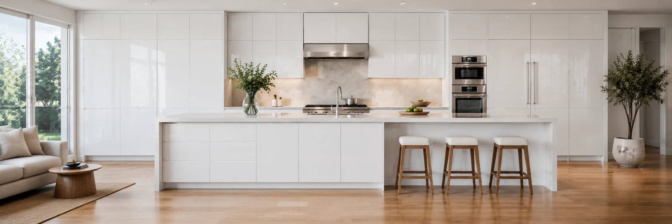 Bright, open-concept modern kitchen featuring white European frameless cabinets, a large matching center island with three wooden bar stools, and built-in stainless steel double ovens.