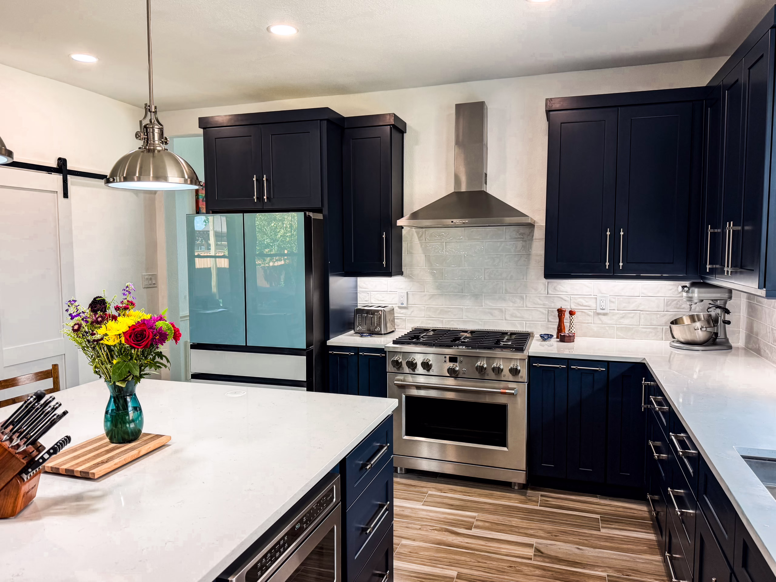 Completed kitchen renovation featuring navy or onyx black shaker cabinets with silver slim handles, quartz countertops, stainless steel range oven with chimney hood, and a center island with fresh flowers
