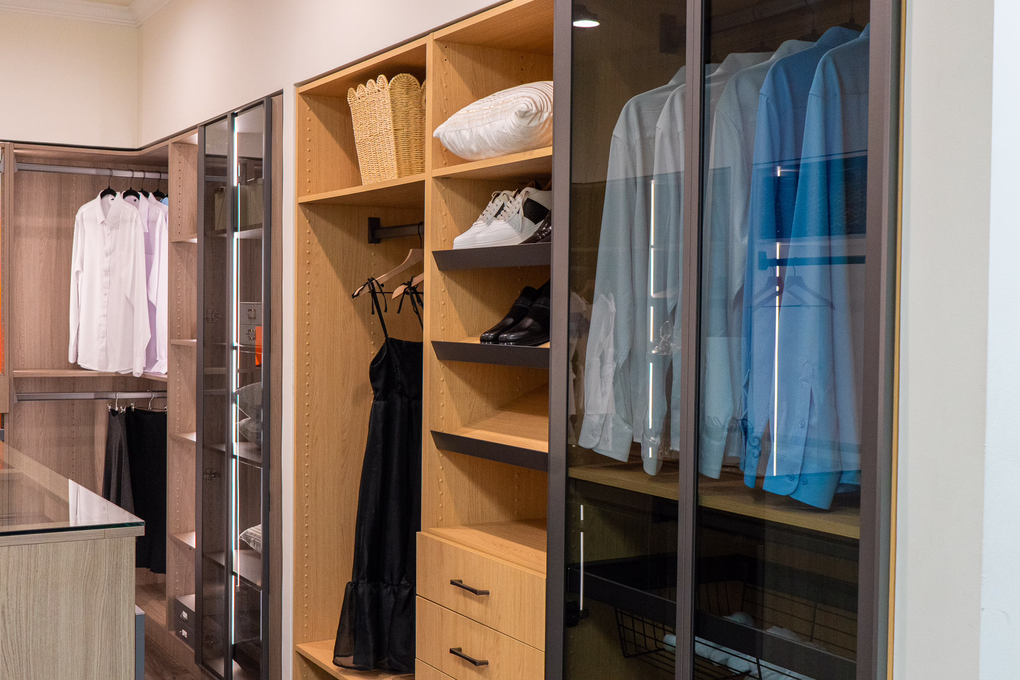 Natural oak walk-in closet display with glass wardrobe doors, open shoe shelving, and built-in LED lighting at Modernform Cabinets and Closets showroom in Carrollton Texas