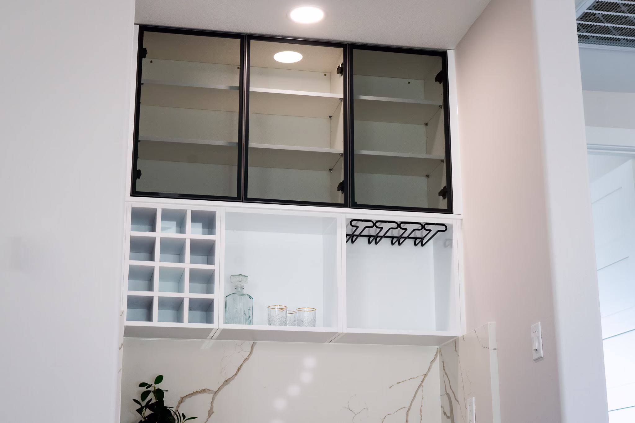 Modern European frameless wall cabinets featuring built-in white grid wine rack storage, glassware holder, and black aluminum-framed glass display doors in a Dallas home.