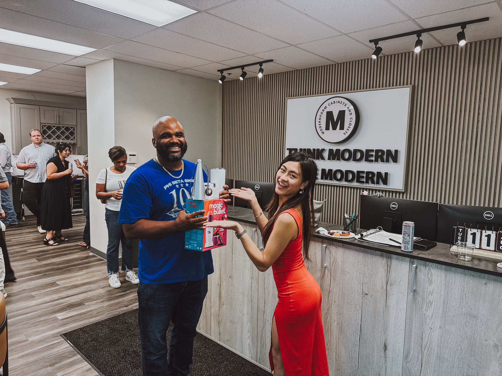 Woman in red dress presenting a boxed prize to a smiling guest during a showroom event at the Modernform Cabinets and Closets reception desk in Carrollton Texas