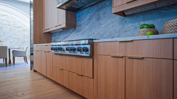 Natural oak European frameless kitchen base cabinets featuring thin gold bar pulls, an integrated stainless steel range cooktop, blue marble backsplash, and under-cabinet lighting.