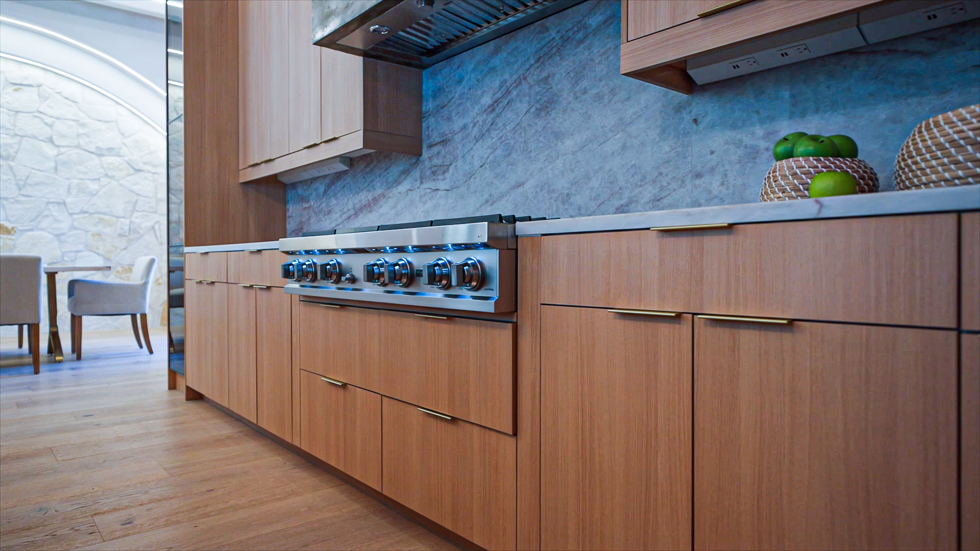 Natural oak European frameless kitchen base cabinets featuring thin gold bar pulls, an integrated stainless steel range cooktop, blue marble backsplash, and under-cabinet lighting.