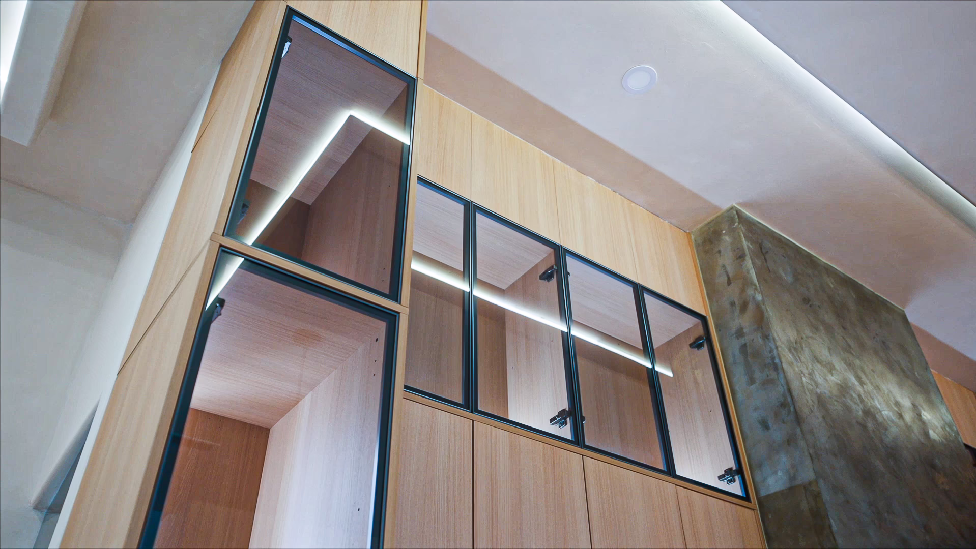 Modern natural oak upper kitchen cabinets with black-framed glass display doors, interior lighting, and sleek hardware mounted against a modern plaster wall.