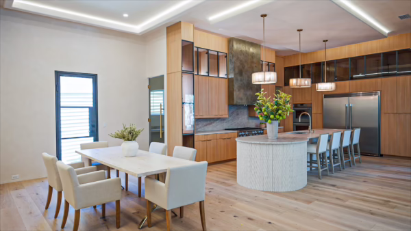 Modern open-concept kitchen featuring natural oak European frameless upper cabinets, a textured curved kitchen island, integrated stainless steel appliances, and an adjacent dining table.