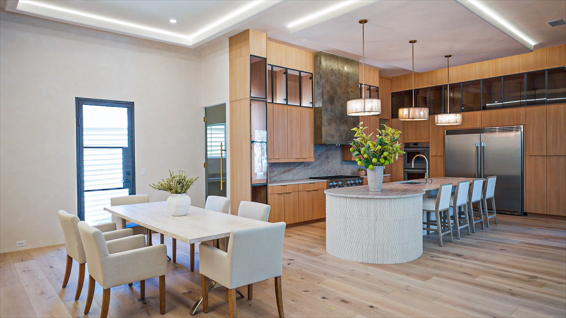 Modern open-concept kitchen featuring natural oak European frameless upper cabinets, a textured curved kitchen island, integrated stainless steel appliances, and an adjacent dining table.
