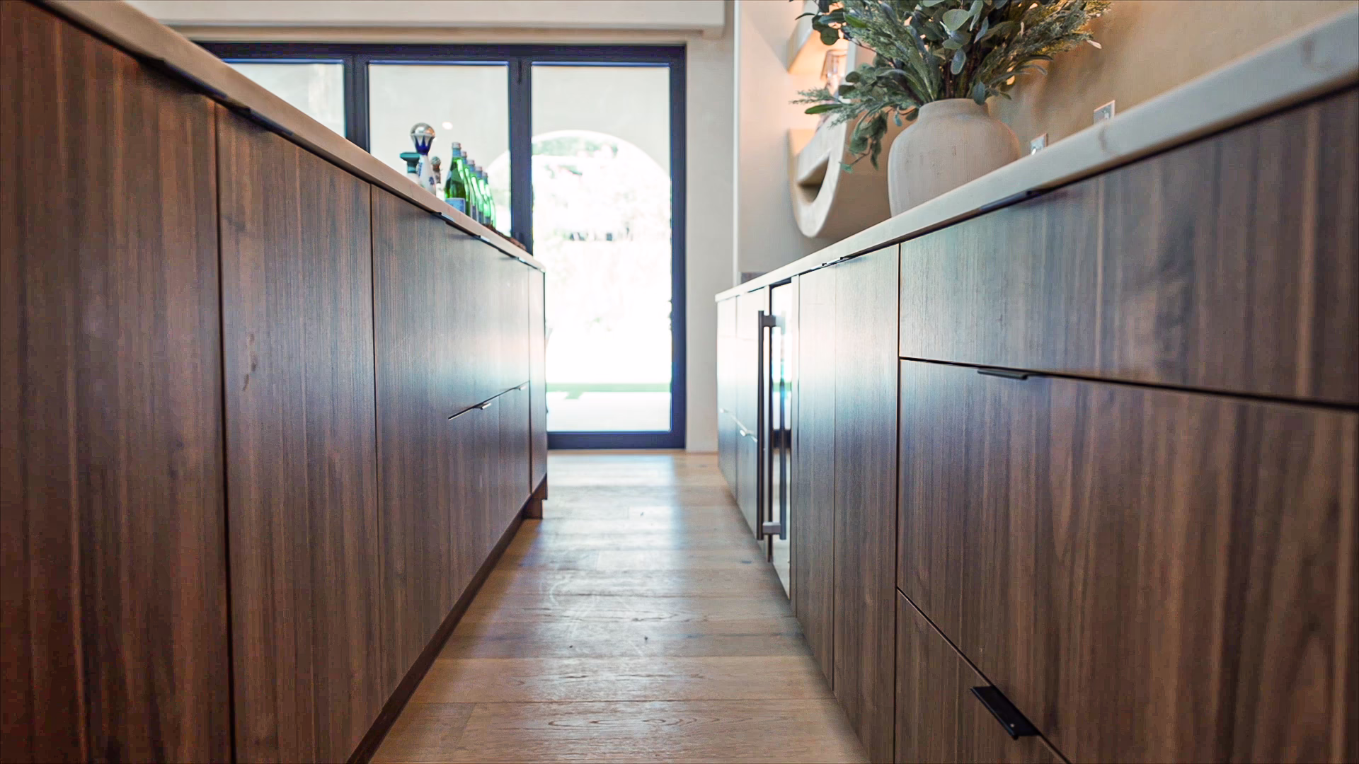 Modern dark walnut European frameless kitchen island and base cabinets with dark slim pull handles leading toward glass patio doors.