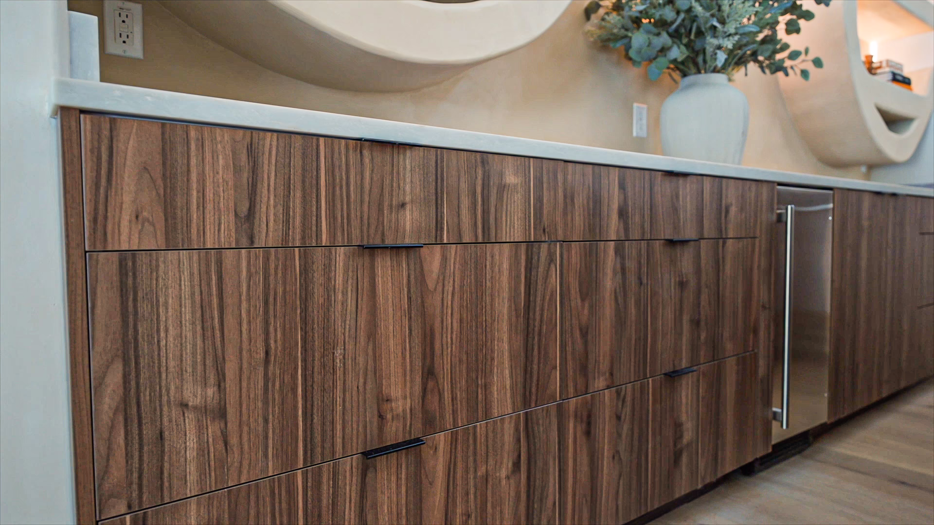 Modern dark walnut European frameless kitchen base cabinets featuring black edge pull handles, light stone countertops, and an integrated stainless steel beverage cooler.