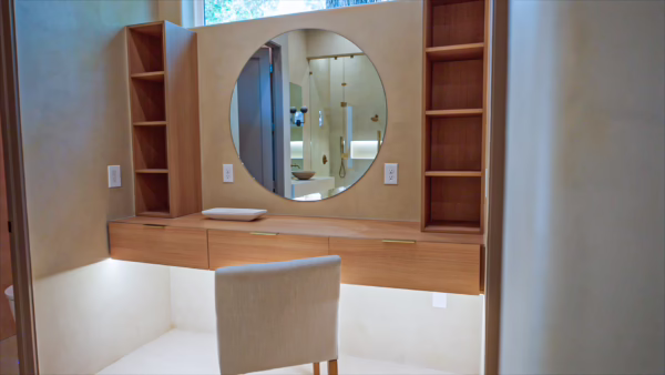 Custom natural oak floating makeup vanity featuring thin brass pull drawers, under-cabinet LED lighting, a large circular wall mirror, and built-in side shelving.
