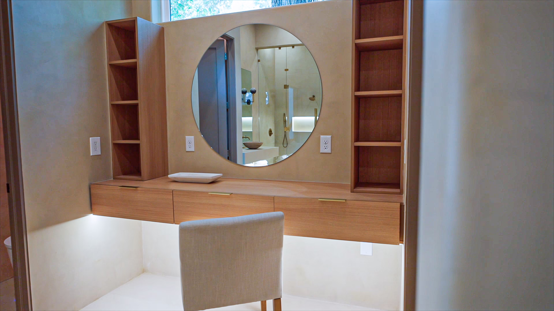 Custom natural oak floating makeup vanity featuring thin brass pull drawers, under-cabinet LED lighting, a large circular wall mirror, and built-in side shelving.
