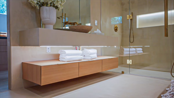 Modern natural oak European frameless floating bathroom vanity featuring under-cabinet LED strip lighting, flat-panel wood drawers, folded white towels, and a glass-enclosed shower background.