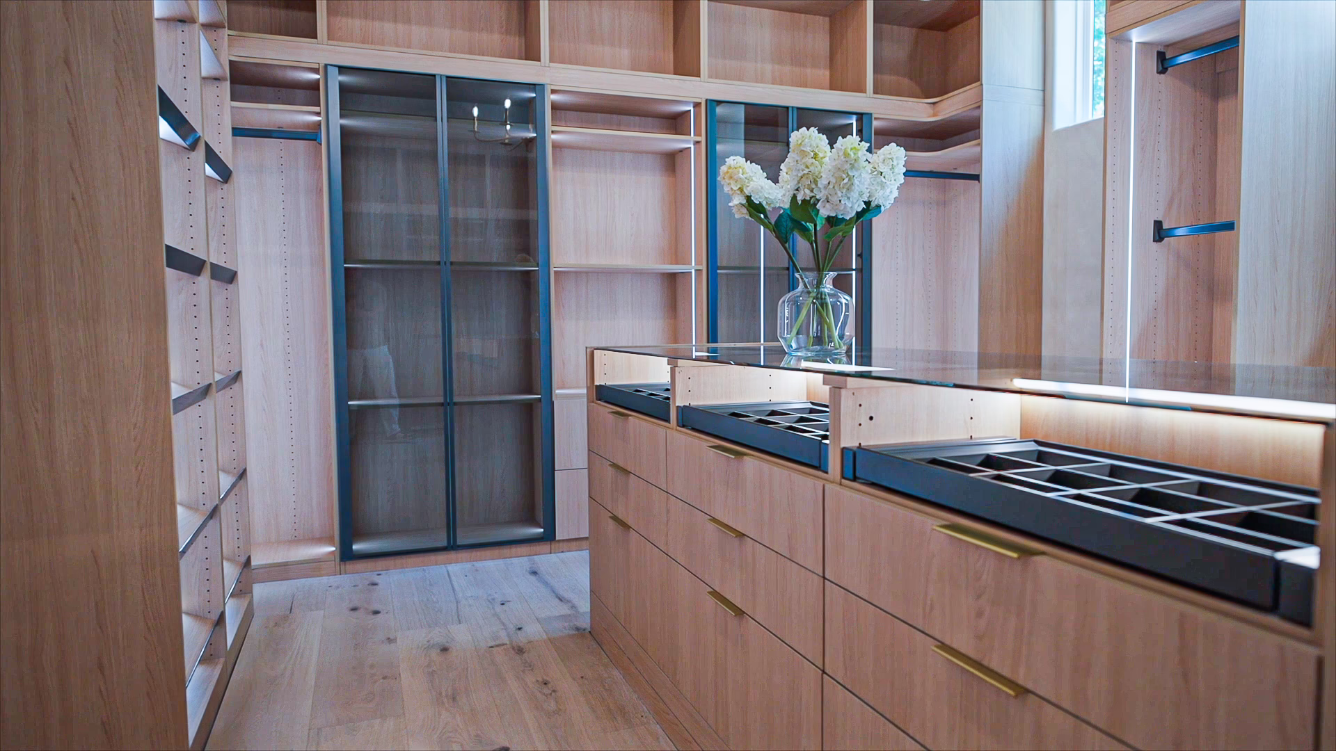 Natural oak walk-in closet island featuring a glass top display drawer, thin gold handles, white flower vase, and glass wardrobe doors with dark blue framing.