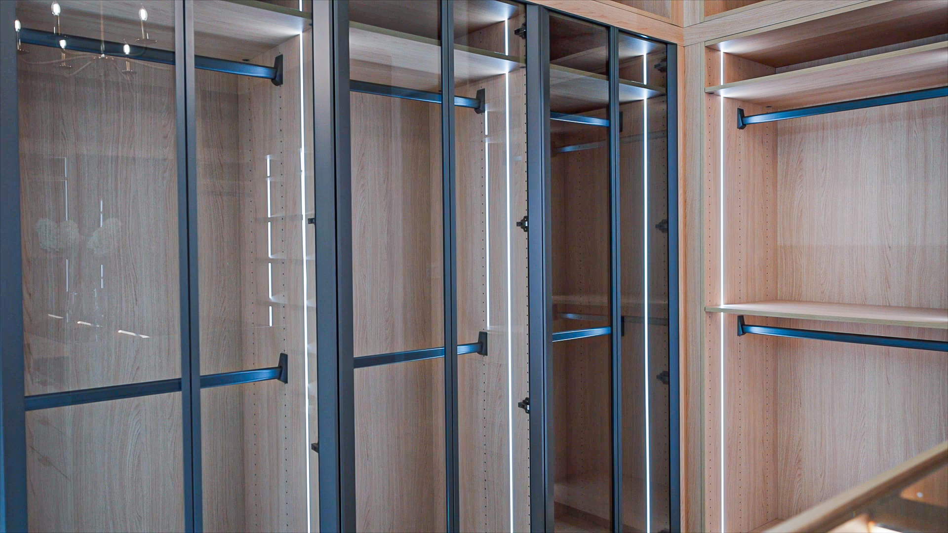 Custom natural oak walk-in closet featuring glass front wardrobe doors with dark frames, open clothing racks, and integrated LED shelf lighting.