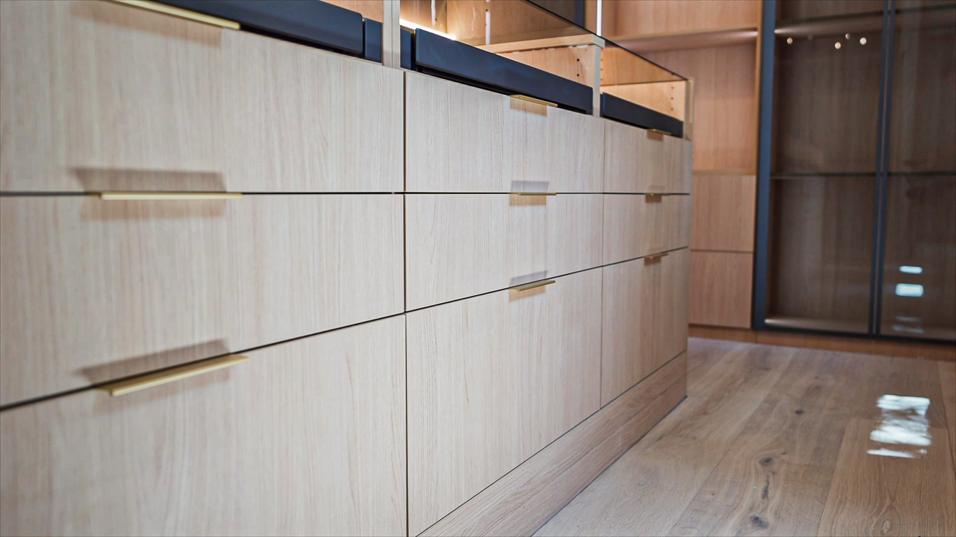 Custom natural oak closet island featuring wide storage drawers with thin brass pull handles, a glass display top, and light hardwood flooring.