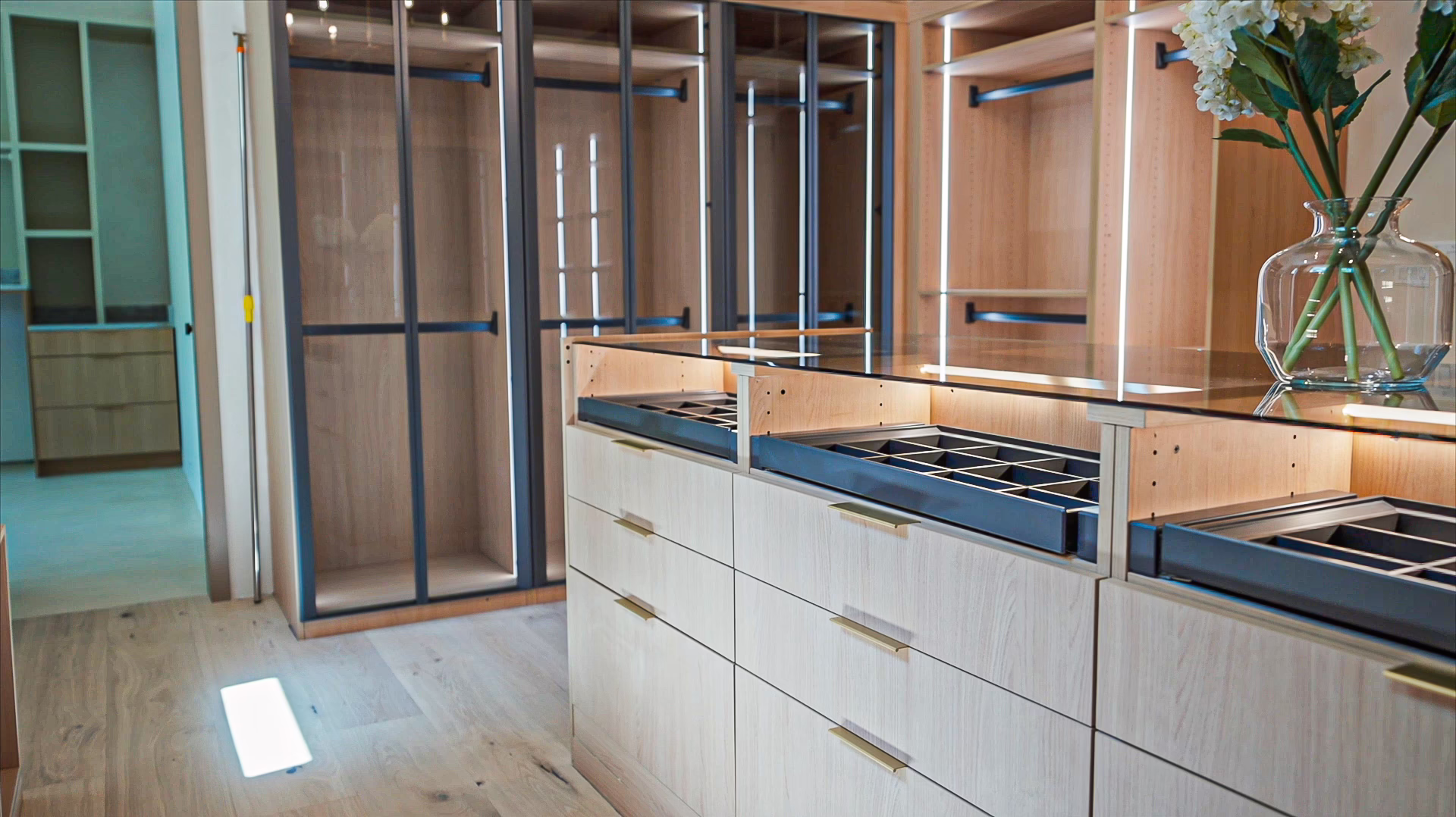 Natural oak custom closet island featuring a glass top view into dark accessory organizer trays and built-in drawer units under soft LED lighting.