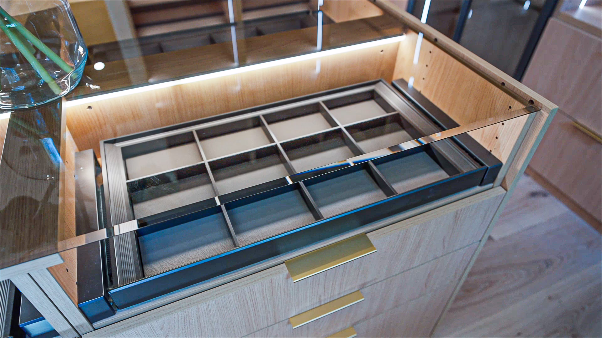 Natural oak custom closet island featuring a glass top view into a dark accessory organizer tray and wide pull-out drawers with modern brass handles.