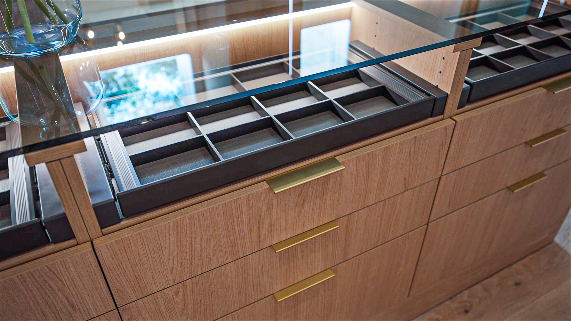 Natural oak custom closet island with gold pull handles and a glass top displaying partitioned accessory storage drawers.