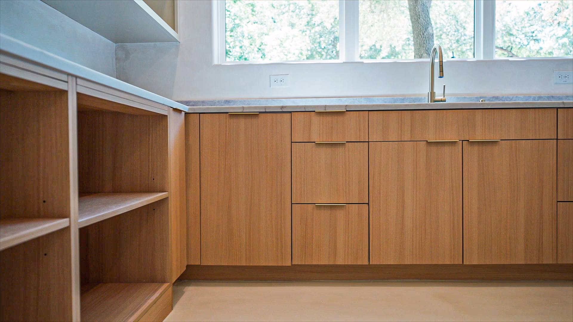 Natural oak European frameless lower kitchen cabinets with thin brass bar pulls, open corner shelving, and light marble countertops below a large window.